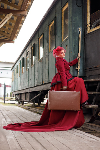 A beautiful girl in a burgundy suit of the last century and a hat with a veil stands with a suitcase in her hands near an old steam locomotive.Vintage portrait of the last century, retro journey.