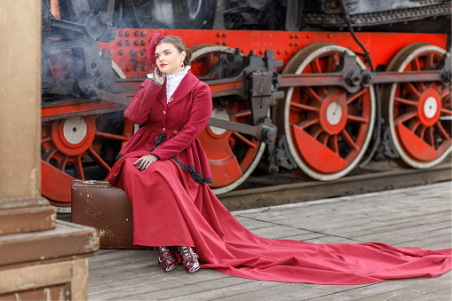 A beautiful girl in a burgundy suit of the last century and a hat with a veil is sitting on a suitcase near an old steam locomotive.Vintage portrait of the last century, retro journey.
