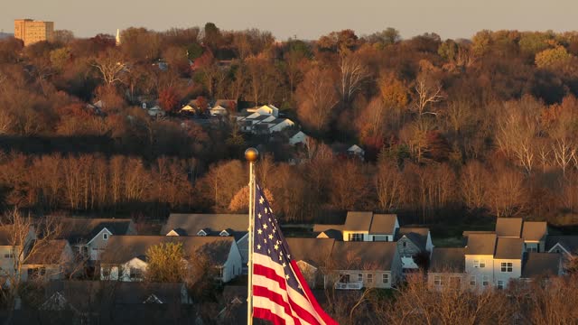 Cinematic shot of American flag waving against American neighborhood amidst bare trees in winter during golden hour sunset. Aerial zoom.