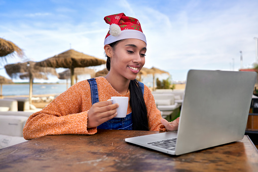 Beautiful Colombian girl works on her digital business from Spain, is having breakfast on a beach while reviewing sales In Christmas with a Santa hat