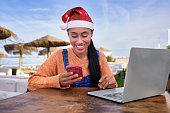 In Christmas a Beautiful Colombian Latin girl who is a digital nomad works outside her home with her laptop and her smart phone in her e-commerce business in Christmas with a Santa hat and smiling seeing the results.