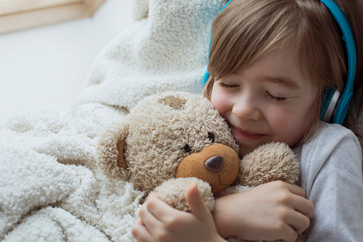Happy cute boy hugs his teddy bear and listens music at his cozy home.