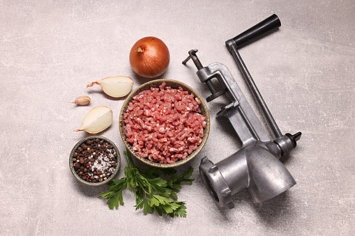 Manual meat grinder with beef mince, peppercorns, onion and parsley on light grey table, flat lay
