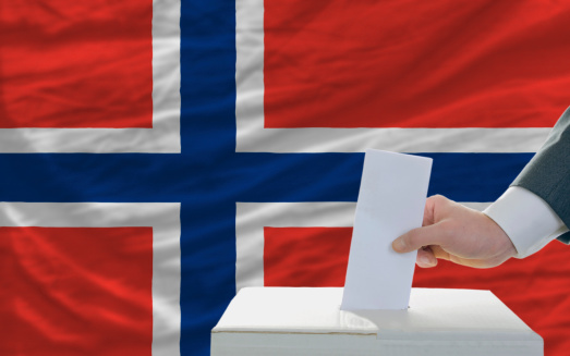 man putting ballot in a box during elections in norway in front of flag