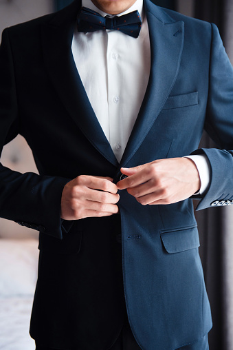 Stylish groom adjusting his black suit while standing at bright room. Young man is getting ready for wedding ceremony. Morning time.