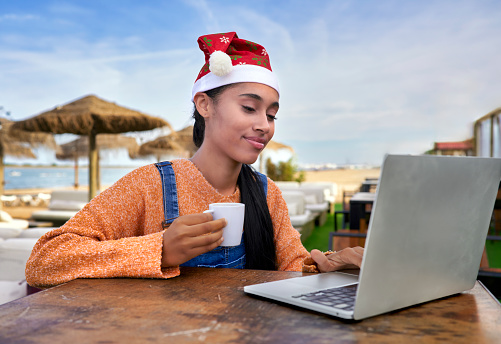 Beautiful Colombian girl works on her digital business from Spain, is having breakfast on a beach while reviewing sales In Christmas with a Santa hat