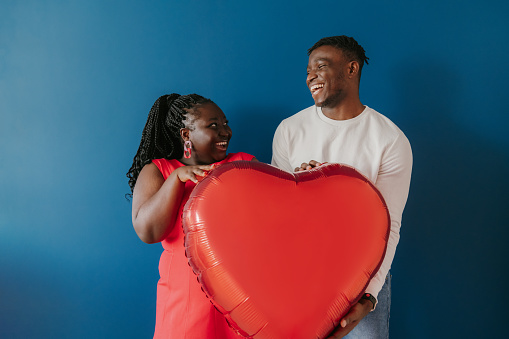 Happy young African couple holding red heart shaped balloon while standing on blue background