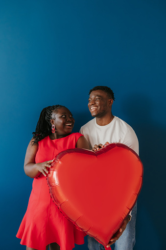 Happy young African couple holding red heart shaped balloon while celebrating Valentines day on blue background