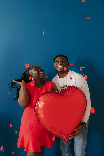 Happy young African couple holding red heart shaped balloon while confetti flying around on blue background