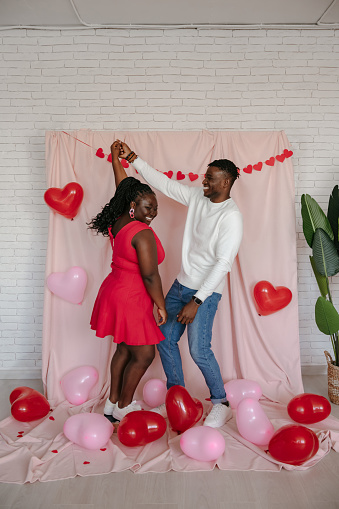 Romantic young African couple dancing on pink fabric background with heart shaped balloons