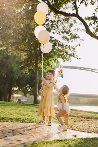 Two little Asian toddler girls are playing balloons and windmill toy in a garden park.