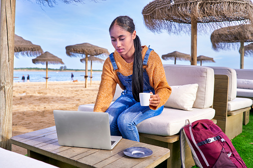 Beautiful Colombian Latin girl who is a digital nomad works in the beach with her laptop in her e-commerce business seeing the results.