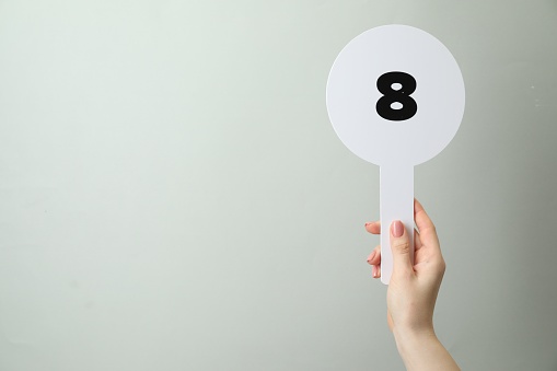 Woman holding auction paddle with number 8 on light grey background, closeup. Space for text