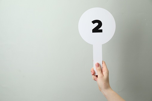 Woman holding auction paddle with number 2 on light grey background, closeup. Space for text