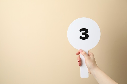 Woman holding auction paddle with number 3 on beige background, closeup. Space for text