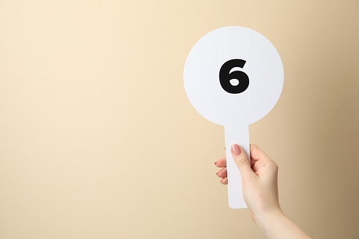 Woman holding auction paddle with number 6 on beige background, closeup. Space for text