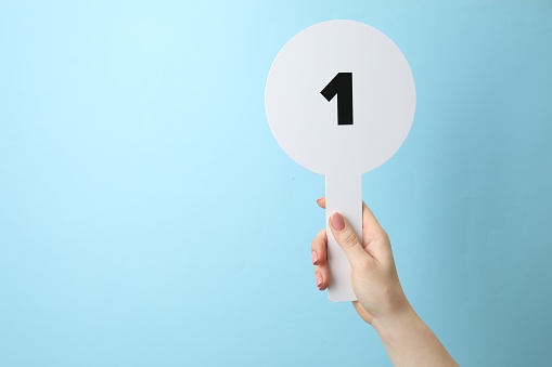 Woman holding auction paddle with number 1 on light blue background, closeup. Space for text