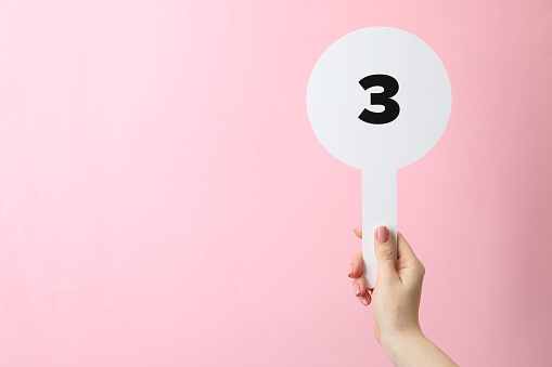 Woman holding auction paddle with number 3 on pink background, closeup. Space for text