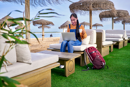 Beautiful Colombian Latin girl who is a digital nomad works in the beach with her laptop in her e-commerce business seeing the results.