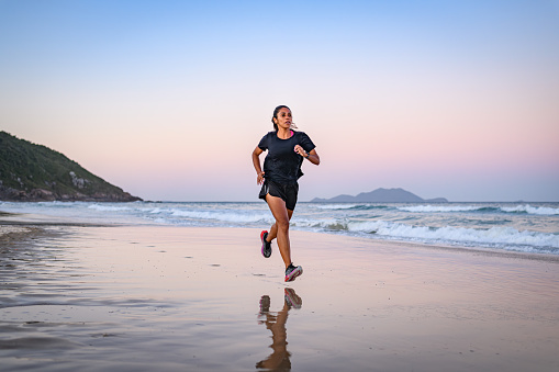 Beautiful black woman athlete performing running training drill on the beach at dusk in summer. Her clear goals and objectives together with her self-discipline lead her to great sporting results at the finish line.