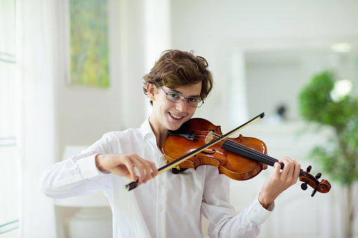 One young adult caucasian man sitting on the chair at home learning to play violin male violinist playing music at his apartment in room alone