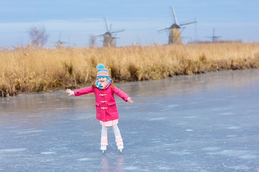 Child ice skating on frozen canal with wind mills and snow in Holland. Little girl with skates on natural ice on cold winter day in the Netherlands. Kids ice skate in snowy Dutch windmill landscape.