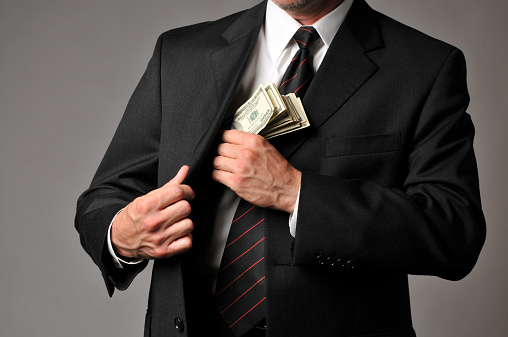 Horizontal studio image of a well dressed businessman putting a stack of $100 bills in his suit coat pocket. This could represent a pay bonus or some kind of embezzlement or corruption. A small amount of post processing noise was added to the gray background.