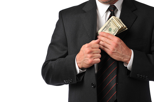 Horizontal studio image of a well dressed businessman putting a stack of $100 bills in his suit coat pocket. This could represent a pay bonus or some kind of embezzlement or corruption. Photograph taken on a white background.