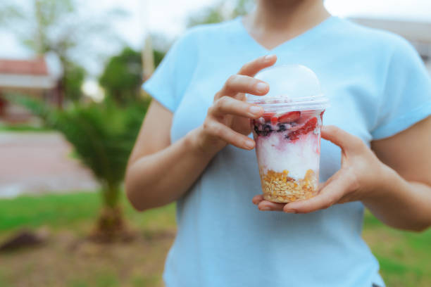 Woman dressed in light blue holding plastic container with fruits and yogurt with hands and copy space