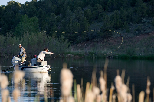 A fisherman in boots stands in the water of a calm river and holds a fishing rod. In front of him is the expanse of a wide river. Trees grow along the banks. View from the back