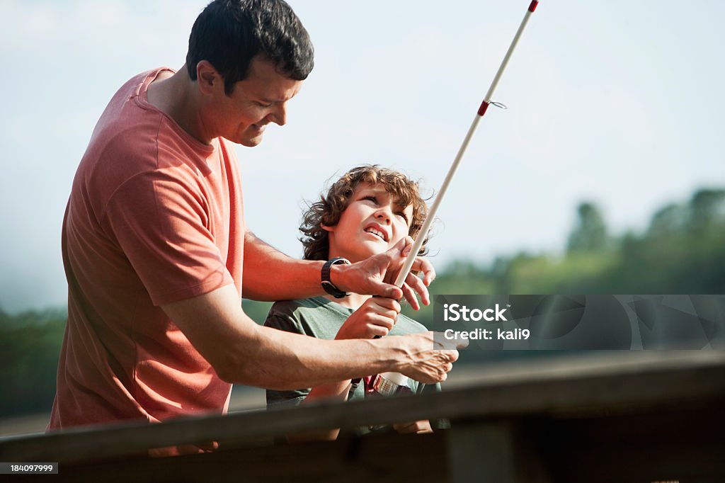 Father and son fishing A father and his 9 year old son on a fishing and camping trip.  He is helping the boy with the rod they are both holding.  They are standing on a wooden foot bridge at the railing.  The main focus is on the face of the child as he looks up at dad.  In the background are clear skies and trees. Fishing Stock Photo