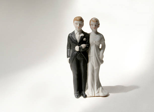 Wedding cake topper figurines of a couple on a soft white background. The antique bride and groom in tuxedo and traditional white gown may represent old-fashioned marriage nuptials or forecasting of anniversaries, eternal love and happiness, breakups, separations, or divorce. The ceramic newlyweds are reception celebration party decorations.
