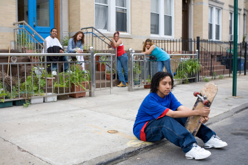 A bored Hispanic teenager sitting on the curb. His friends in the background. Selective focus. Low income houses in the background.