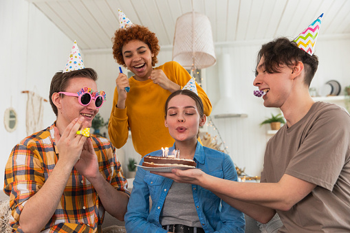 Make a wish. Woman wearing party cap blowing out burning candles on birthday cake. Happy Birthday party. Group of friends wishes girl happy birthday. People celebrating birthday with party at home