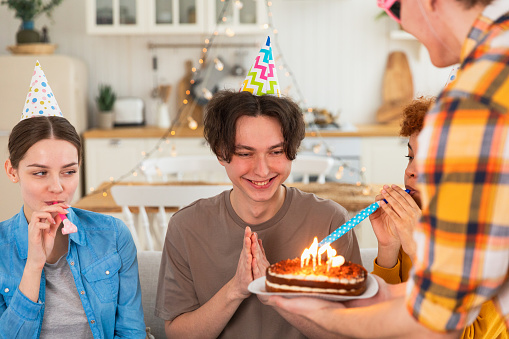 Make a wish. Man wearing party cap blowing out burning candles on birthday cake. Happy Birthday party. Group of friends wishes guy happy birthday. People celebrating birthday with party at home