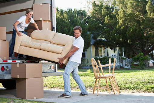 A young couple lifts a tan couch to move it in or out of the back of a moving truck.  A young woman with blond hair pulled away from her face is standing inside the truck holding the left end of the couch.  The woman wears a white top and dark blue jeans.  A young man with brown hair stands outside the truck holding the right end of the couch.  The man is wearing a white shirt and light blue jeans.  Moving boxes and sit inside and outside the truck.  Two chairs sit on a sidewalk outside the truck.  A yellow house can be seen in the background.