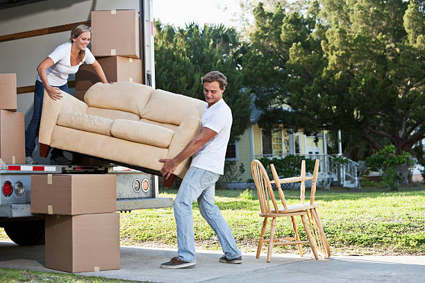 Young couple moving house A young couple lifts a tan couch to move it in or out of the back of a moving truck.  A young woman with blond hair pulled away from her face is standing inside the truck holding the left end of the couch.  The woman wears a white top and dark blue jeans.  A young man with brown hair stands outside the truck holding the right end of the couch.  The man is wearing a white shirt and light blue jeans.  Moving boxes and sit inside and outside the truck.  Two chairs sit on a sidewalk outside the truck.  A yellow house can be seen in the background. moving van stock pictures, royalty-free photos & images