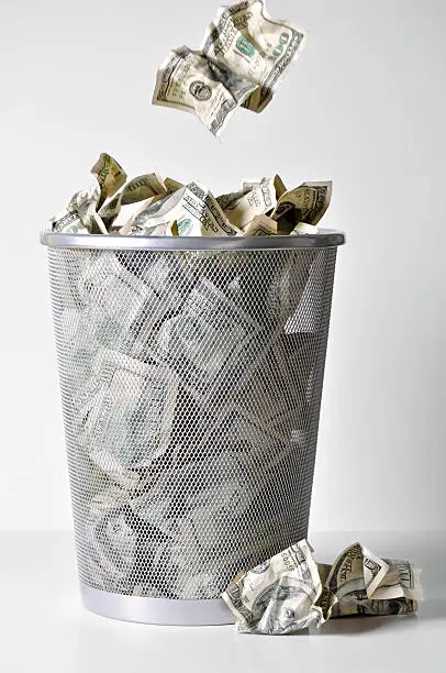 Conceptual image expressing the value of money decreasing or a metaphor for throwing your money away. A trashcan is full of wadded up $100 and $50 bills. Another $100 is in motion toward the trashcan. A small amount of grain has been added to the background in post processing.