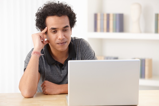 Young afro-american man in casual clothes sitting at desk at home, using laptop computer, touching head, looking at screen, smiling.