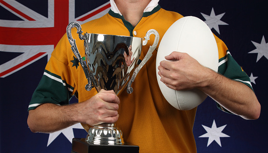 A rugby player holds a trophy and a ball in front of an Australian flag