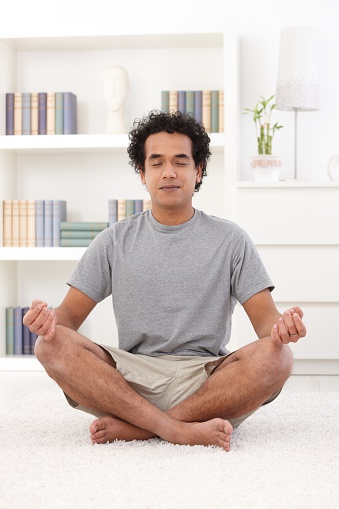 Young afro-american man in casual clothes, sitting in lotus position at home in living room, eyes closed, smiling.
