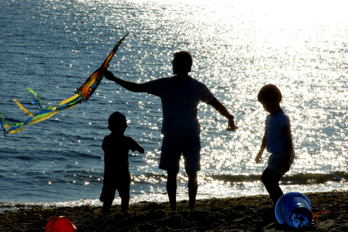 Father and sons flying a kite and playing in the sand at the beach.  Sunset reflecting off of the ocean.