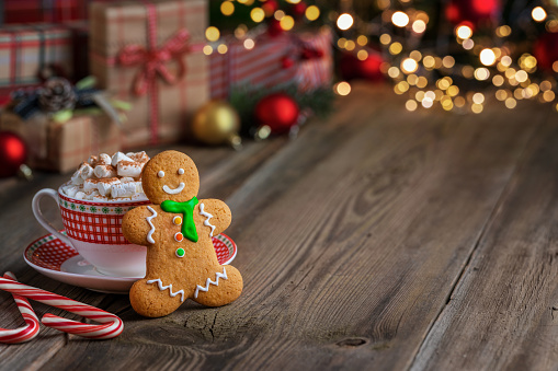 Homemade gingerbread man cookie and cup of hot chocolate with marshmallows and candy cane on the wooden table. Defocused lights and Christmas decorations and gifts on background. Copy space.