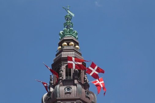 Copper spire on top of the Danish parliament which is situated in Christiansborg castle in the middle of Copenhagen. The current Christiansborg is build in a neo-baroque style. The construction took place from 1906 to 1928 after a fire had destructed the former castle. Notice the brown colour of new copper and the green colour of the old. The Danish flag is called Dannebrog and is one of the oldest in the world. This tailed shape is only legal when used by permission of the king / queen.