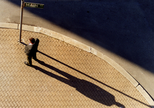 A bird's eye view of a man standing on a sidewalk next to a street sign in the late afternoon sun.