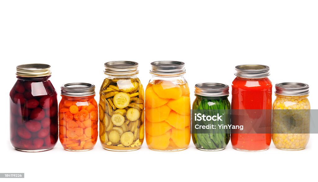 Canning Jars of Canned, Pickled Vegetable Food Preserved for Storage Horizontal straight-on view of a row of seven different canned vegetables and fruit, lined up in glass canning jars. The variety of vegetables that are sauced, pickled, or sliced include carrots, green beans, tomato sauce, corn, dill pickles, beats, and peaches, prepared for preserved storage. Isolated on a white background. Jar Stock Photo