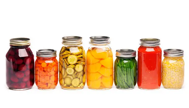 Horizontal straight-on view of a row of seven different canned vegetables and fruit, lined up in glass canning jars. The variety of vegetables that are sauced, pickled, or sliced include carrots, green beans, tomato sauce, corn, dill pickles, beats, and peaches, prepared for preserved storage. Isolated on a white background.