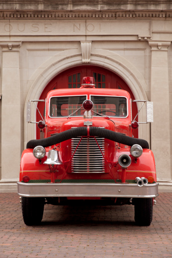 This is a photo of a front view of an old antique firetruck in front of a retro fire station. The focus is on the front of the truck and falls off in the background.Click on the links below to view lightboxes.