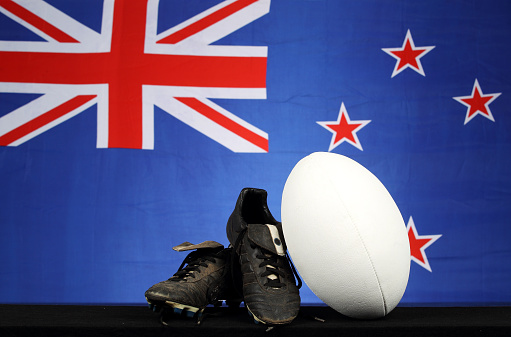 A pair of rugby boots and a ball in front of a New Zealand flag
