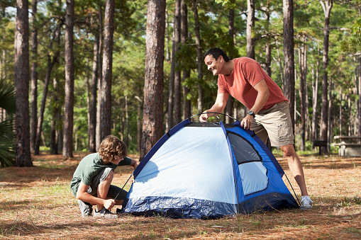 A man with his 9 year old son on a family camping trip, setting up a small tent in a clearing in the woods on a sunny day.  The man is standing on one side of the blue tent.  The boy is kneeling on the other side.  There are trees in the background.